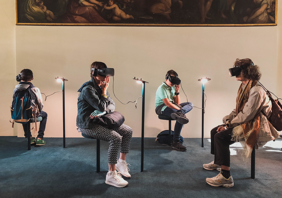 Four persons in a museum are wearing VR-Goggles and sit in front of a painting