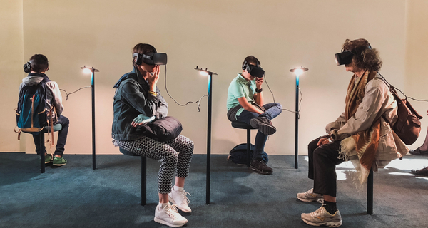 Four persons in a museum are wearing VR-Goggles and sit in front of a painting.