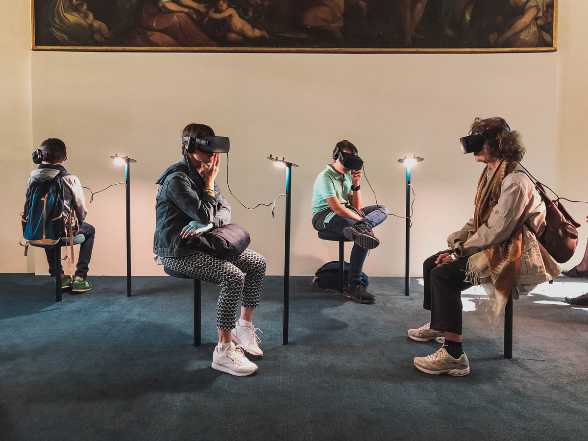 Four persons in a museum are wearing VR-Goggles and sit in front of a painting.
