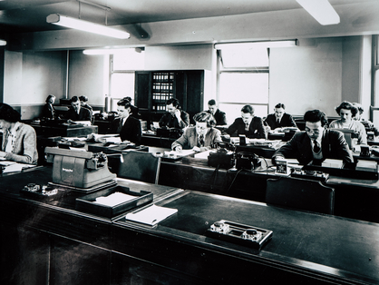 Old black-and-white photography of multiple people working at desks in the office.